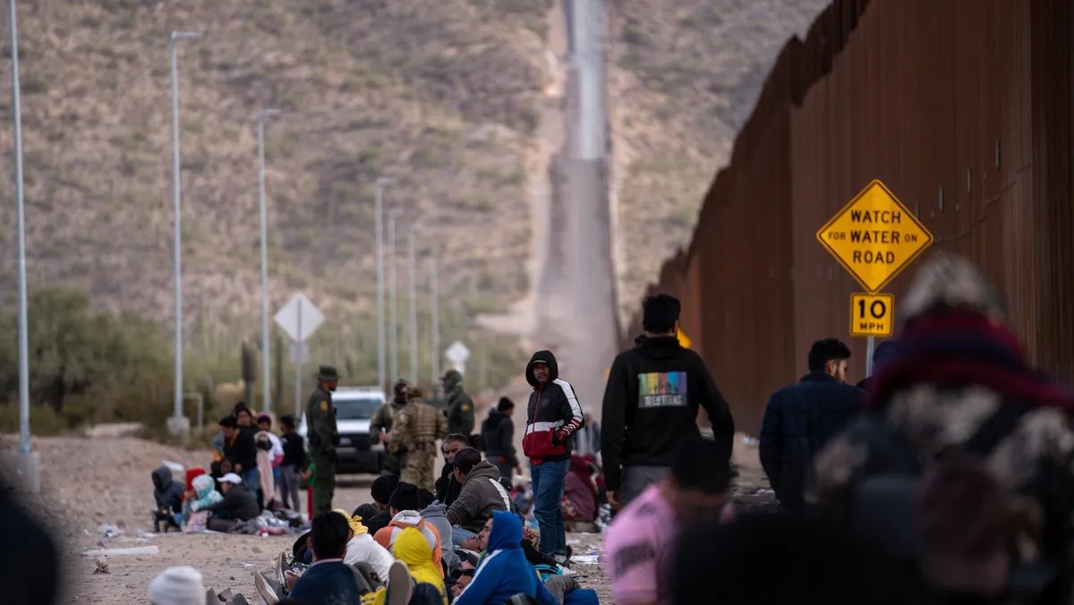 A somber group of asylum seekers waits patiently near a border fence, their faces etched with uncertainty as they hope for entry into the United States under the contested metering policy.