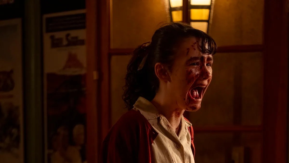 A shadowy figure reminiscent of Pennywise looming over a distressed depiction of Derry, Maine, with a cloudy, ominous sky above, hinting at the sinister nature of Welcome To Derry.