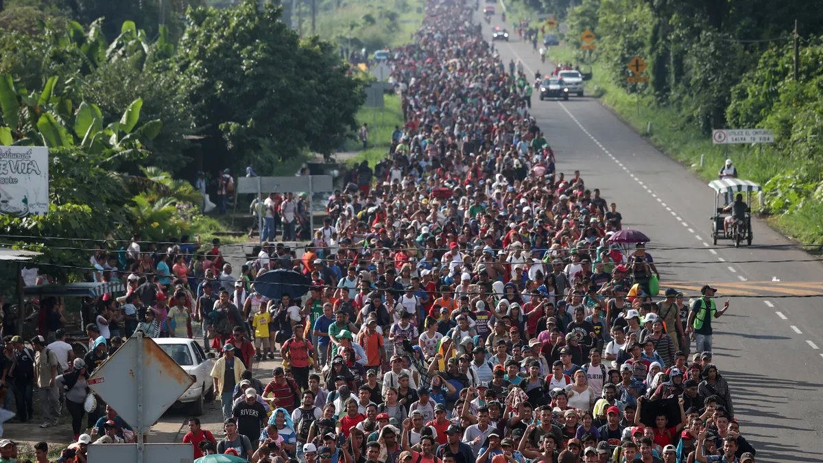 A large group of migrants walking on a road in Mexico, many carrying backpacks and belongings, suggesting they are part of a migrant caravan.