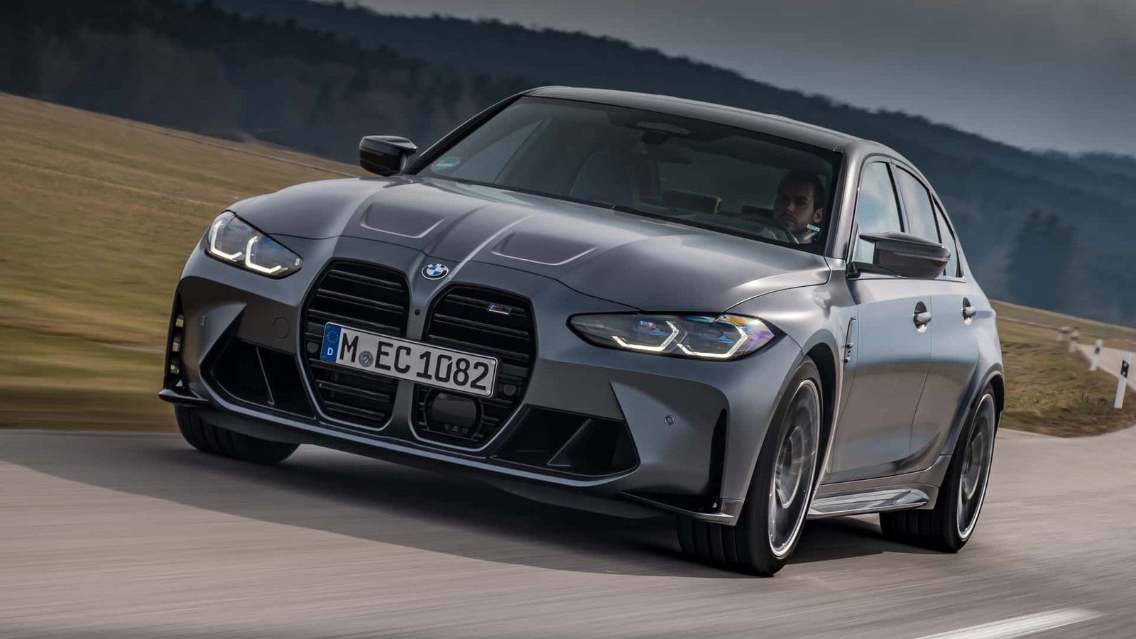 A sleek, modern BMW M Competition model, possibly an M3 or M4, driving dynamically on a track with a clear sky.