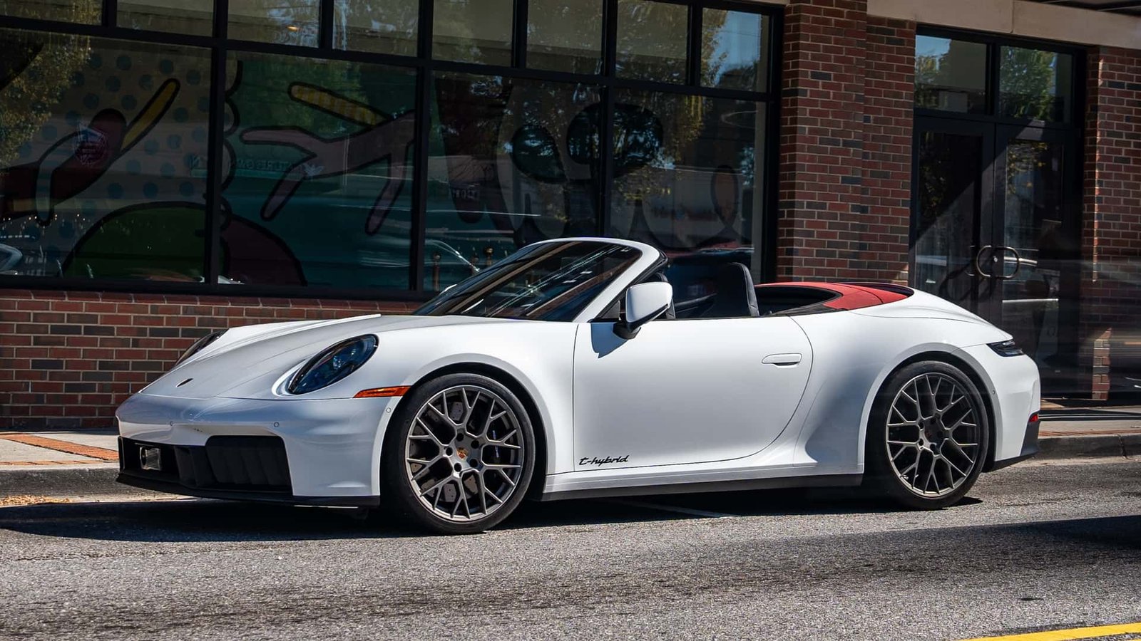 A sleek, grey Porsche 911 Carrera GTS with a red stripe, parked on a race track with a mountainous backdrop, showcasing its performance-oriented design.