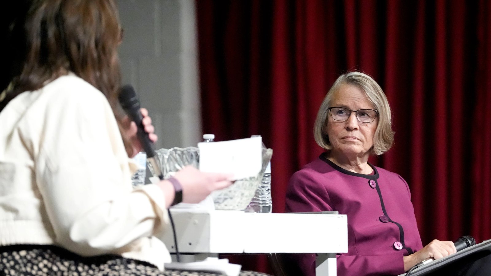 A concerned woman in a town hall audience, raising her hand to ask a question, her expression serious and focused.