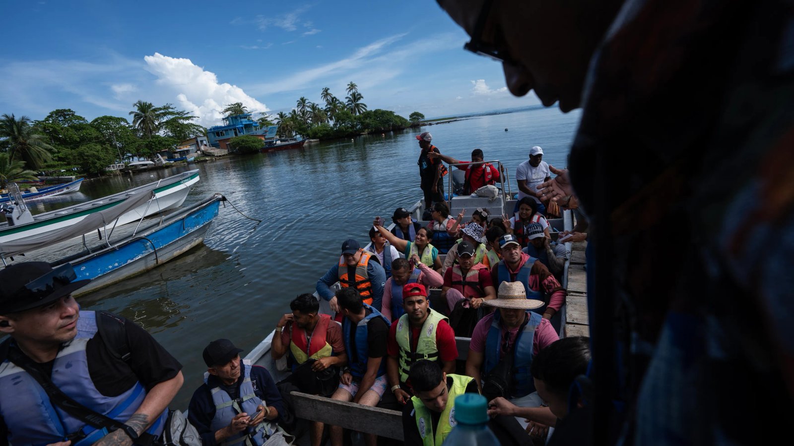 A small group of migrants looking forlorn on a boat, with lush Panamanian greenery in the background, signifying their journey south.