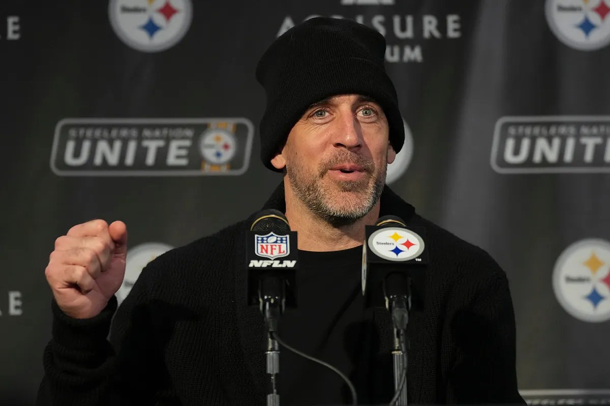 Aaron Rodgers in a Jets uniform, looking thoughtful on the field with a stadium background