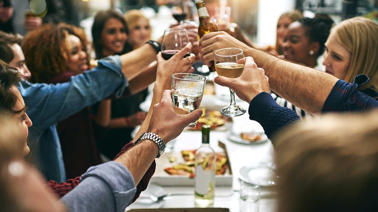 A group of young adults laughing and enjoying a meal at a communal table in a trendy restaurant.