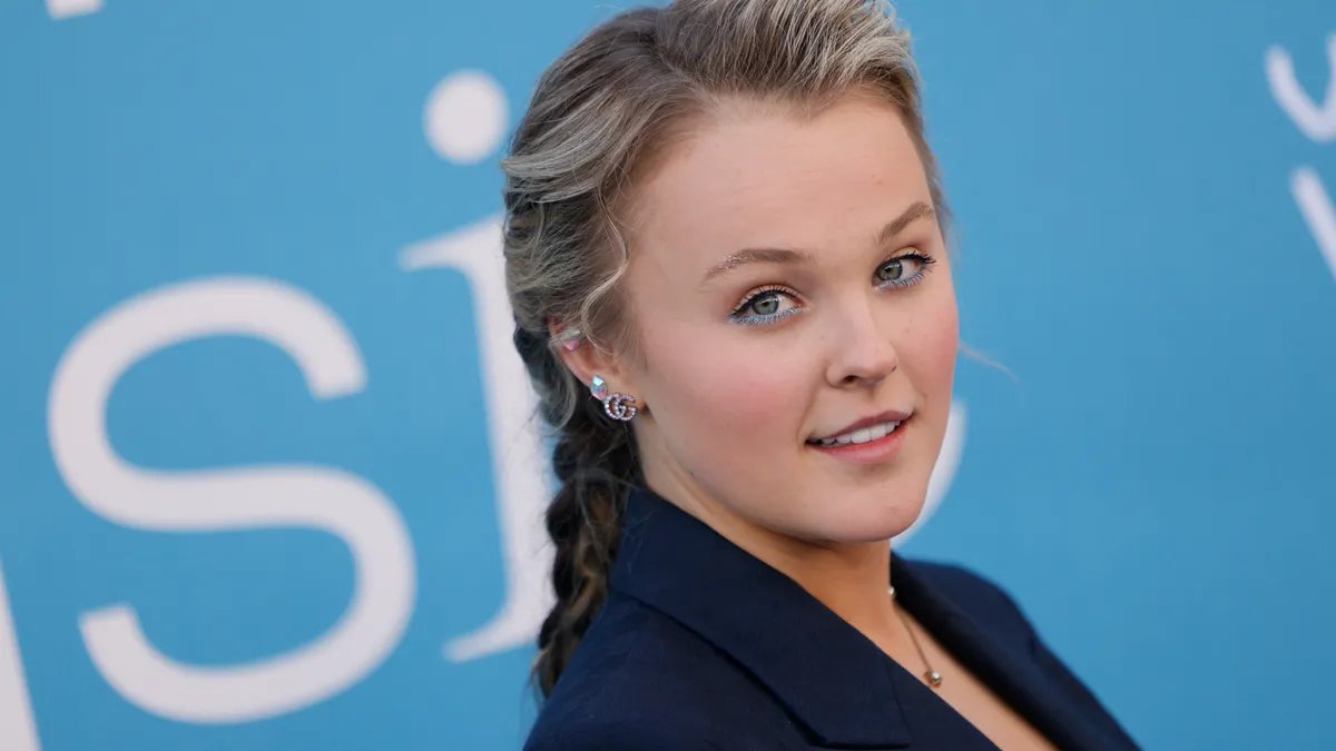 JoJo Siwa in hospital bed, looking pale but giving a thumbs up.
